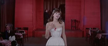 Movie still from “A Woman Is a Woman” (1961), directed by Jean-Luc Godard – A woman in a white dress is standing in front of a red wall; Medium shot, Low angle