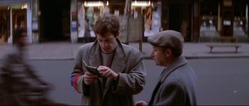 Movie still from “A Woman Is a Woman” (1961), directed by Jean-Luc Godard – A man looking at a cell phone while standing next to another man; Medium shot, Over the shoulder angle