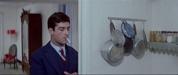 Movie still from “A Woman Is a Woman” (1961), directed by Jean-Luc Godard – A man smoking a cigarette next to a pot hanging on the wall; Medium shot, Over the shoulder angle
