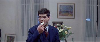 Movie still from “A Woman Is a Woman” (1961), directed by Jean-Luc Godard – A man in a suit and tie eating a piece of food; Medium shot, Over the shoulder angle