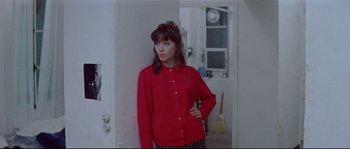 Movie still from “A Woman Is a Woman” (1961), directed by Jean-Luc Godard – A woman standing in a room wearing a red jacket; Medium shot, Over the shoulder angle