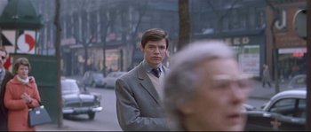 Movie still from “A Woman Is a Woman” (1961), directed by Jean-Luc Godard – A man in a suit and tie standing on the street; Medium shot, Over the shoulder angle