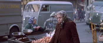 Movie still from “A Woman Is a Woman” (1961), directed by Jean-Luc Godard – An older woman standing in front of a bunch of carrots; Medium shot, High angle