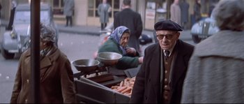 Movie still from “A Woman Is a Woman” (1961), directed by Jean-Luc Godard – An older man and woman are standing in front of a hot dog cart; Medium shot, Low angle