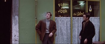 Movie still from “A Woman Is a Woman” (1961), directed by Jean-Luc Godard – A man standing in front of a building smoking a cigarette; Medium shot, Low angle