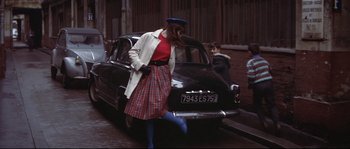 Movie still from “A Woman Is a Woman” (1961), directed by Jean-Luc Godard – A woman in a plaid skirt is standing next to an old car; Wide shot, High angle