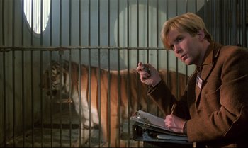 Movie still from “A Zed & Two Noughts” (1985), directed by Peter Greenaway – A man holding a cell phone in front of a tiger cage; Medium shot, Low angle