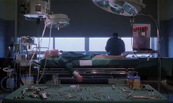 Movie still from “A Zed & Two Noughts” (1985), directed by Peter Greenaway – A man laying on a hospital bed in a room; Wide shot, Low angle