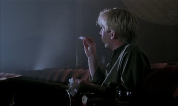 Movie still from “A Zed & Two Noughts” (1985), directed by Peter Greenaway – A man sitting in front of a couch holding a piece of paper; Medium shot, Low angle