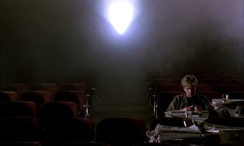 Movie still from “A Zed & Two Noughts” (1985), directed by Peter Greenaway – A person sitting in a theater with a light shining on them; Wide shot, High angle