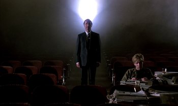 Movie still from “A Zed & Two Noughts” (1985), directed by Peter Greenaway – A man in a suit standing in front of a bunch of papers; Wide shot, Low angle