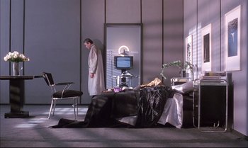 Movie still from “A Zed & Two Noughts” (1985), directed by Peter Greenaway – A man standing in front of a bed in a room; Wide shot, High angle