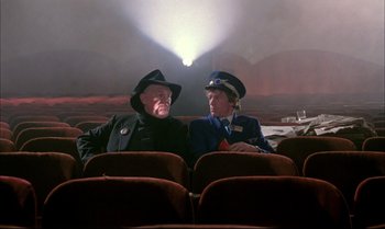 Movie still from “A Zed & Two Noughts” (1985), directed by Peter Greenaway – Two people sitting in a theater with a projector in the background; Wide shot, Low angle