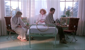 Movie still from “A Zed & Two Noughts” (1985), directed by Peter Greenaway – A group of people sitting on a bed in a room; Wide shot, High angle