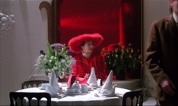 Movie still from “A Zed & Two Noughts” (1985), directed by Peter Greenaway – A woman sitting at a table with a red hat on; Medium shot, Low angle