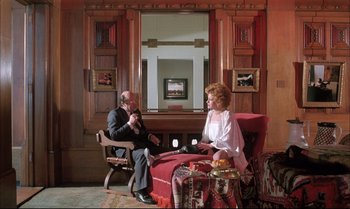 Movie still from “A Zed & Two Noughts” (1985), directed by Peter Greenaway – A man and a woman sitting on a red couch in front of a mirror; Wide shot, High angle