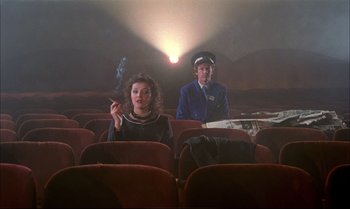 Movie still from “A Zed & Two Noughts” (1985), directed by Peter Greenaway – A woman sitting in a movie theater while a man stands in the background smoking a cigarette; Wide shot, Low angle