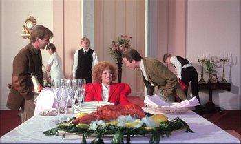 Movie still from “A Zed & Two Noughts” (1985), directed by Peter Greenaway – A woman in a red jacket sitting at a dinner table; Wide shot, Over the shoulder angle
