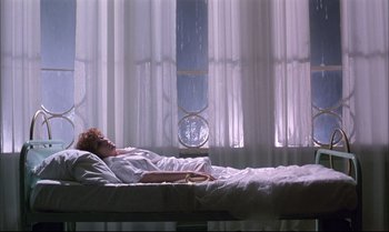 Movie still from “A Zed & Two Noughts” (1985), directed by Peter Greenaway – A woman laying in a bed in front of a window; Wide shot, Low angle
