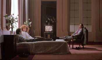 Movie still from “A Zed & Two Noughts” (1985), directed by Peter Greenaway – A man and a woman sitting on a bed in front of a tv; Wide shot, High angle