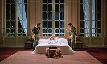 Movie still from “A Zed & Two Noughts” (1985), directed by Peter Greenaway – Three people standing around a woman laying on a bed; Wide shot, Low angle