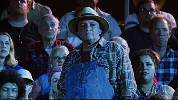Movie still from “A.I. Artificial Intelligence” (2001), directed by Steven Spielberg – An older man wearing a hat and overalls stands in front of a group of older people; Medium shot, Low angle