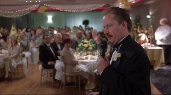 Movie still from “About Schmidt” (2002), directed by Alexander Payne – A man in a suit is holding a microphone in front of a group of people; Medium shot, Low angle