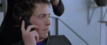 Movie still from “About a Boy” (2002), directed by Chris Weitz – A man talking on a cell phone while a hair stylist is using a blow dryer on his head; Close Up shot, Over the shoulder angle