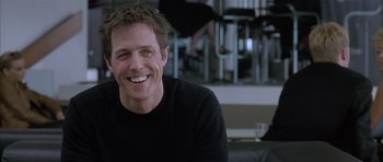 Movie still from “About a Boy” (2002), directed by Chris Weitz – A man smiling for the camera while sitting in a chair; Close Up shot, Over the shoulder angle