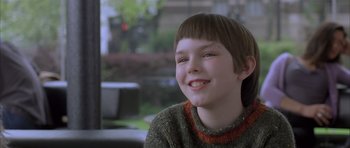 Movie still from “About a Boy” (2002), directed by Chris Weitz – A young boy smiling for the camera in front of a window; Close Up shot, Over the shoulder angle