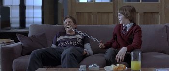 Movie still from “About a Boy” (2002), directed by Chris Weitz – Two people sitting on a couch and one of the people is holding a chain to his mouth; Medium shot, Over the shoulder angle