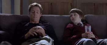 Movie still from “About a Boy” (2002), directed by Chris Weitz – A man smoking a cigarette while a girl sits next to him on a couch; Medium shot, Over the shoulder angle