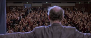 Movie still from “About a Boy” (2002), directed by Chris Weitz – A crowd of people sitting and standing in front of a speaker; Wide shot, Over the shoulder angle