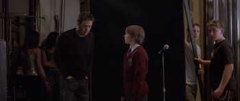 Movie still from “About a Boy” (2002), directed by Chris Weitz – A man standing next to a boy in front of a microphone; Medium shot, Over the shoulder angle
