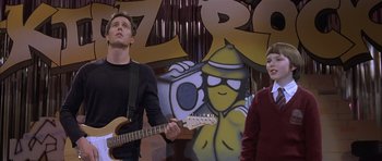 Movie still from “About a Boy” (2002), directed by Chris Weitz – A man and a boy are playing guitar in front of a mural; Medium shot, Over the shoulder angle