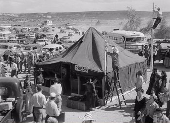 Movie still from “Ace in the Hole” (1951), directed by Billy Wilder – An old black and white photo of a man standing on a ladder next to a tent; Extreme Wide shot, High angle