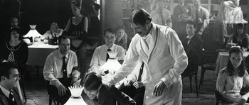 Movie still from “Adam Resurrected” (2008), directed by Paul Schrader – A man in a suit and tie standing in front of a table; Medium shot, Over the shoulder angle