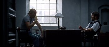 Movie still from “Adam's Apples” (2005), directed by Anders Thomas Jensen – A man sitting in front of a desk eating a donut; Medium shot, Over the shoulder angle