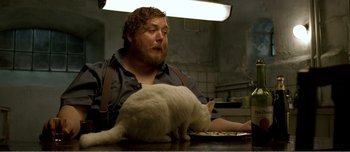 Movie still from “Adam's Apples” (2005), directed by Anders Thomas Jensen – A man sitting at a table next to a white cat; Medium shot, Low angle