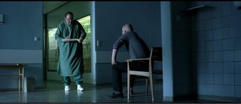 Movie still from “Adam's Apples” (2005), directed by Anders Thomas Jensen – A man sitting on a chair next to another man in a room; Wide shot, High angle