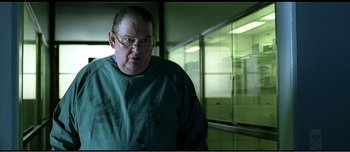Movie still from “Adam's Apples” (2005), directed by Anders Thomas Jensen – A man wearing a green shirt and glasses in a room with glass walls; Medium shot, Over the shoulder angle