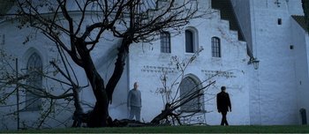 Movie still from “Adam's Apples” (2005), directed by Anders Thomas Jensen – A man standing in front of a building next to a tree; Extreme Wide shot, Low angle