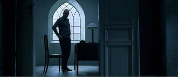 Movie still from “Adam's Apples” (2005), directed by Anders Thomas Jensen – A man standing next to a window in a room; Wide shot, Low angle