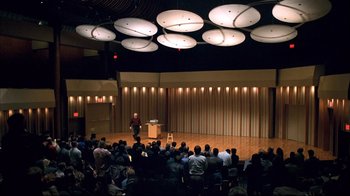 Movie still from “Adaptation.” (2002), directed by Spike Jonze – An audience is sitting in front of a man on a stage; Extreme Wide shot, High angle