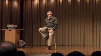 Movie still from “Adaptation.” (2002), directed by Spike Jonze – An older man sitting on top of a wooden stool on a stage; Wide shot, Over the shoulder angle