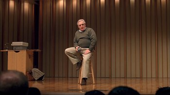 Movie still from “Adaptation.” (2002), directed by Spike Jonze – An older man sitting on top of a wooden stool on a stage; Wide shot, Over the shoulder angle
