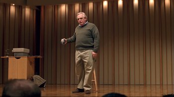 Movie still from “Adaptation.” (2002), directed by Spike Jonze – An older man standing on a stage holding an object; Medium shot, Over the shoulder angle