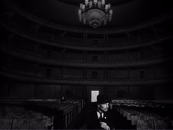 Movie still from “Address Unknown” (1944), directed by William Cameron Menzies – A man in a hat and suit sitting in an empty auditorium; Wide shot, High angle