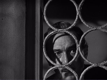 Movie still from “Address Unknown” (1944), directed by William Cameron Menzies – A black and white photo of a man looking through a fence; Close Up shot, Low angle
