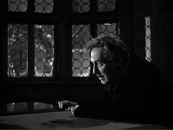 Movie still from “Address Unknown” (1944), directed by William Cameron Menzies – A black and white photo of a man leaning on a table; Close Up shot, Low angle